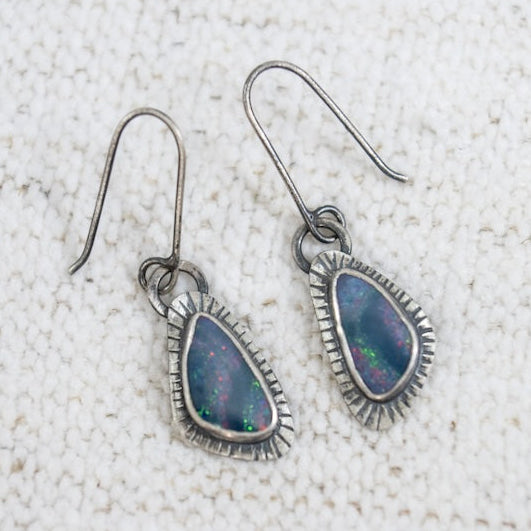 A pair of silver dangle earrings with irregular teardrop-shaped iridescent opal stones, displayed on a textured white woven fabric background.