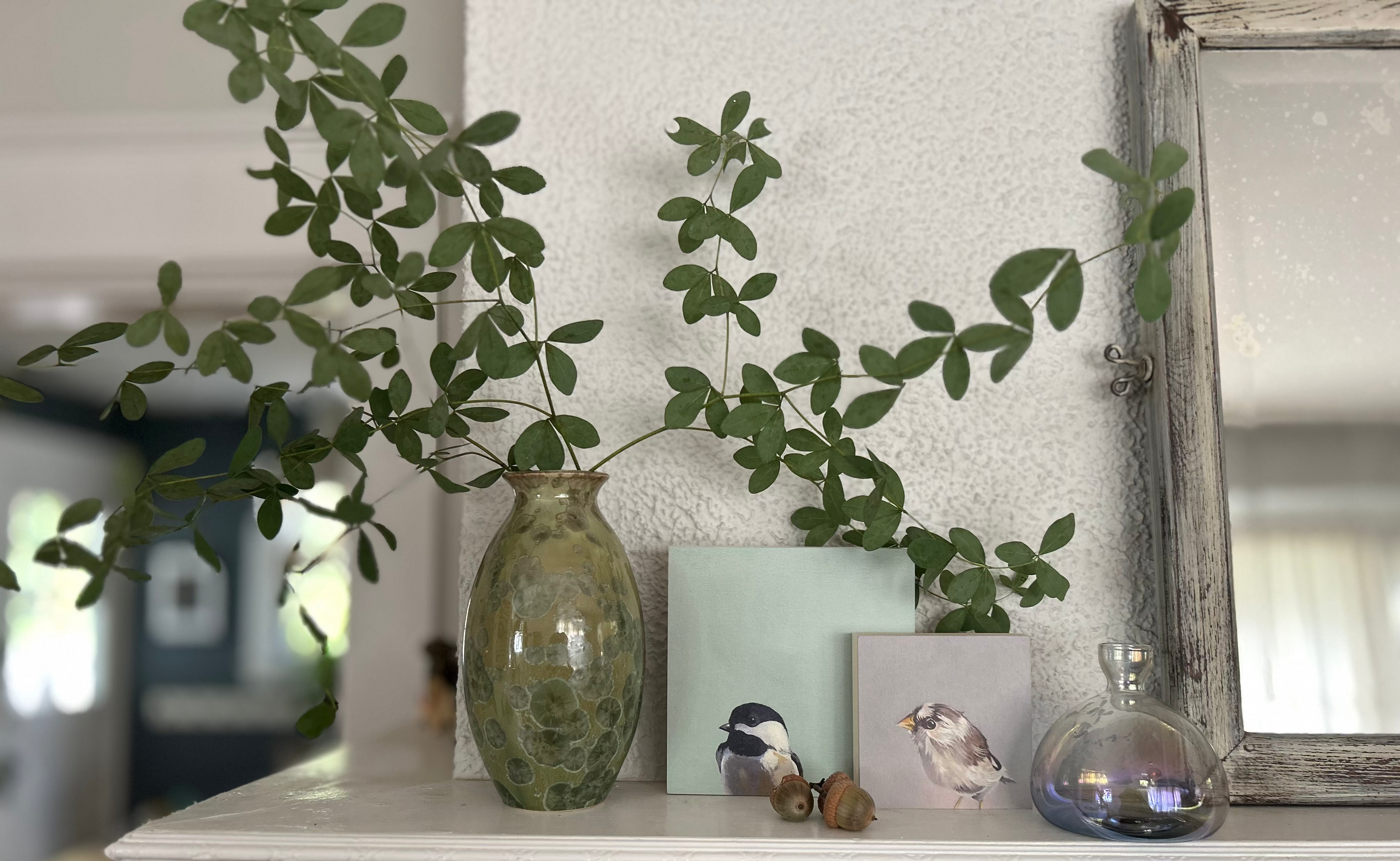 A rustic green glass vase filled with leafy green branches, styled on a white fireplace mantel alongside bird art prints, small acorns, and a small glass bottle.